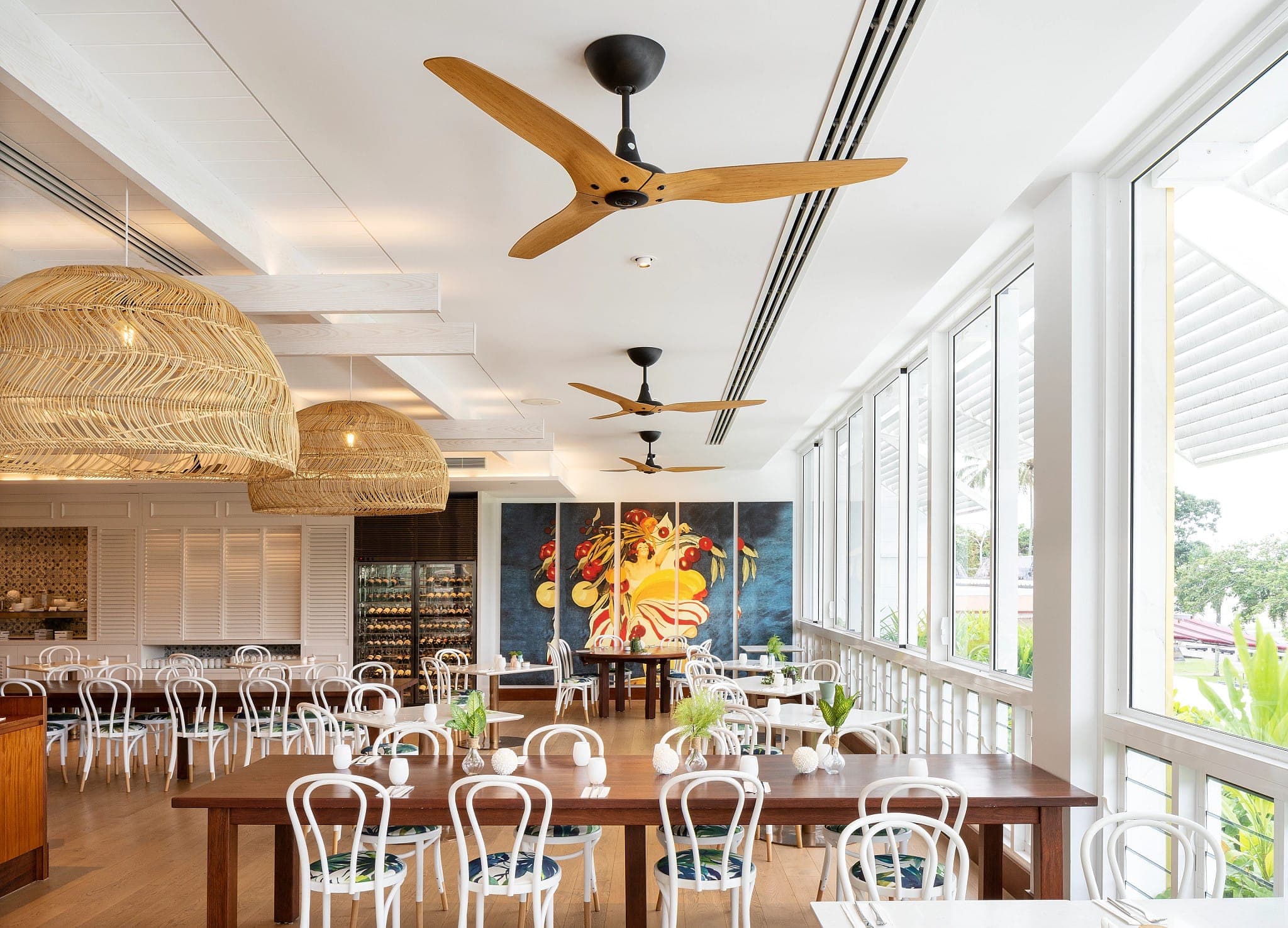 Comedor de restaurante con mesas de madera, sillas blancas de bistró, lámparas colgantes de fibra natural, un mural con frutas y ventanales al jardín; del plafón blanco cuelgan tres ventiladores de tres aspas color madera con base negra.