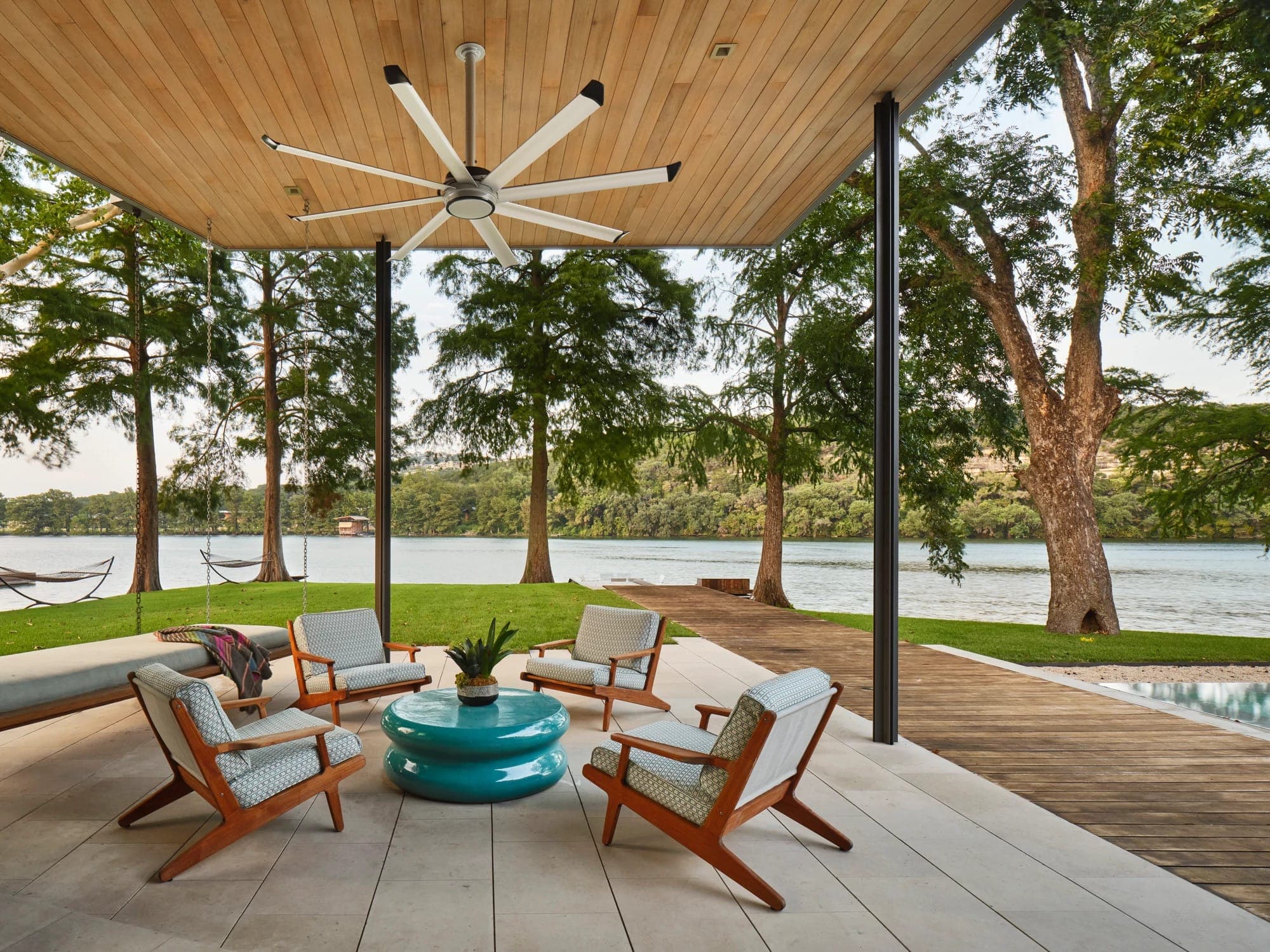 Terraza techada con plafón de madera frente a un lago: cuatro sillones de madera con cojines estampados alrededor de una mesa turquesa y una cama colgante; en el techo, un ventilador de ocho aspas plateadas con puntas negras.