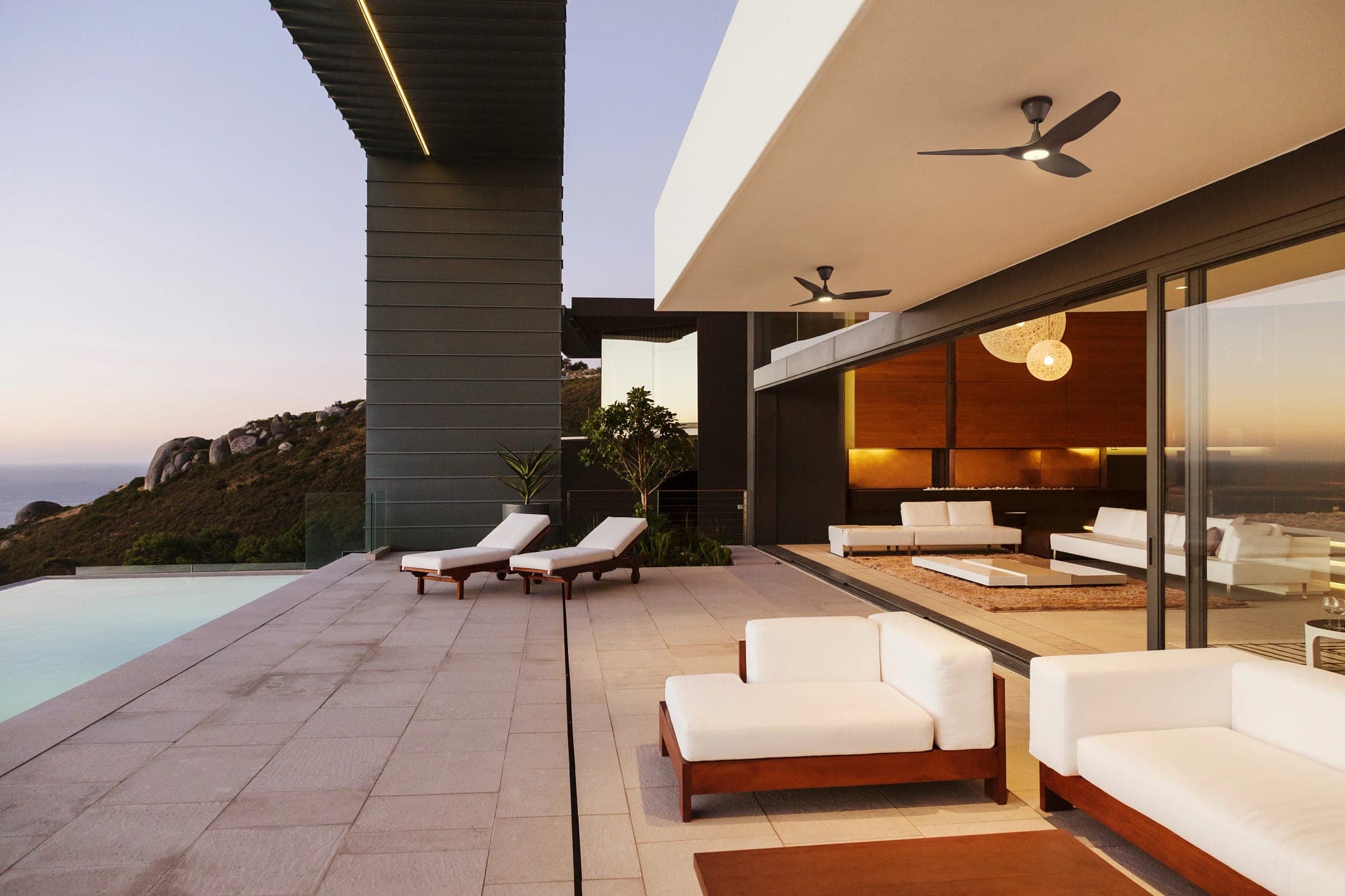 Terraza de una casa moderna al atardecer, con alberca, dos camastros, sillones exteriores blancos con base de madera y la sala interior a la vista; bajo el alero, dos ventiladores negros de tres aspas con la luz encendida; al fondo, un cerro rocoso y el mar.