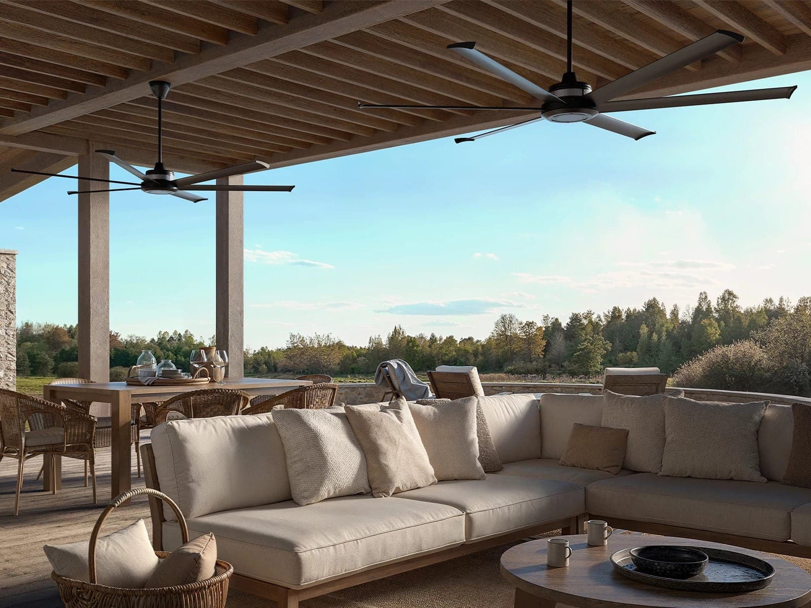 Terraza techada con vigas de madera, sala modular color arena con cojines, mesa de centro redonda de madera y un comedor con sillas de mimbre; en el techo, dos ventiladores de cuerpo negro y aspas largas grises; al fondo, una pradera y un bosque.