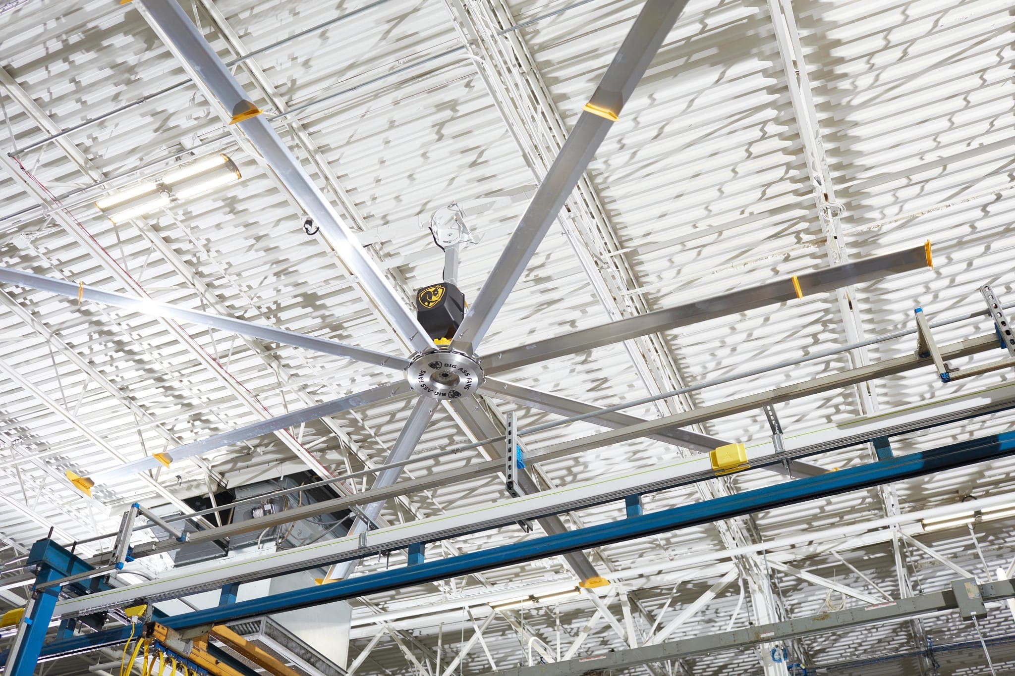 Vista desde abajo de un ventilador industrial de aspas de aluminio con piezas amarillas, bajo la estructura blanca de una cubierta; el cubo lleva la leyenda BIG ASS FANS y, sobre él, una carcasa negra muestra el emblema de la marca; abajo, vigas azules de una grúa.