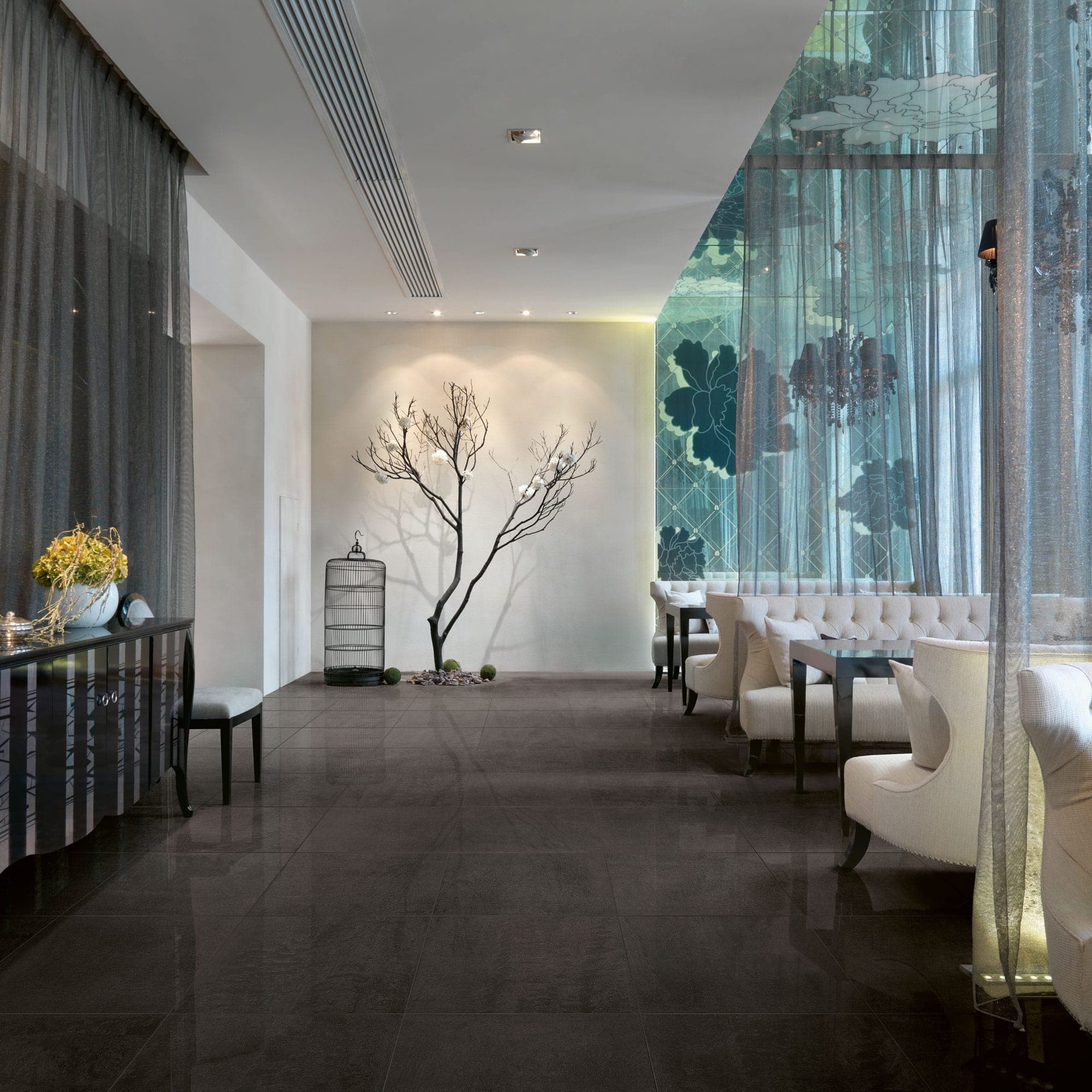 Salón con piso negro pulido que refleja la luz, cortinas translúcidas, una rama decorativa y una jaula frente a un muro blanco y, a la derecha, sillones capitonados blancos junto a mesas.