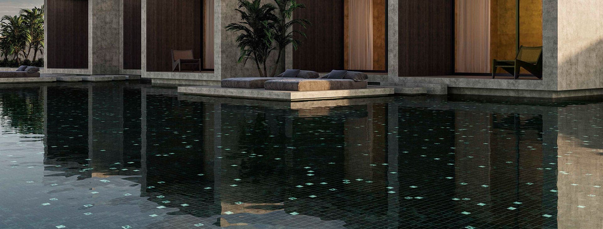 Alberca de fondo oscuro con teselas sueltas de color verde claro, frente a un edificio de concreto con muros de madera oscura; sobre una plataforma en el agua, dos camastros con cojines grises.