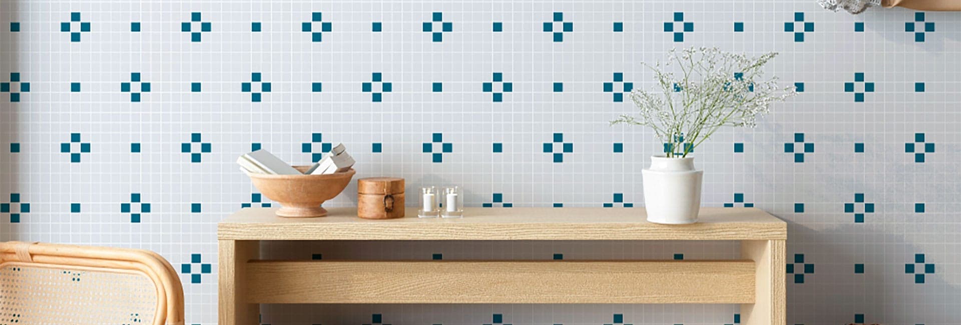 Muro de mosaico cuadrado blanco con un patrón de cruces y puntos en azul petróleo, detrás de una consola de madera clara con dos velas en vasos de vidrio y un florero blanco con flores pequeñas.