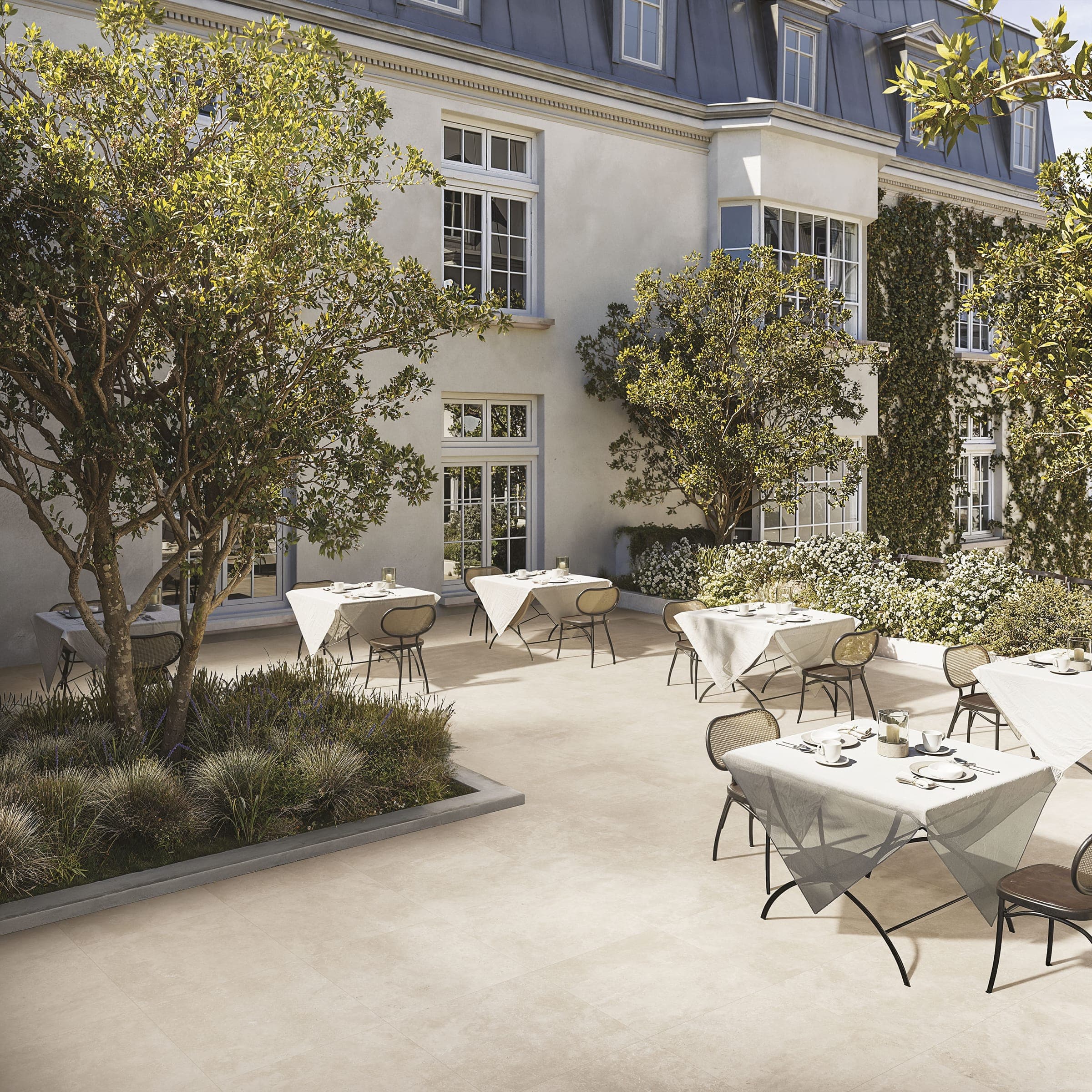 Terraza de restaurante frente a un edificio clásico de fachada blanca con mansarda; mesas con mantel blanco, sillas de rejilla, árboles y una jardinera con pastos sobre un piso exterior de piezas color marfil.