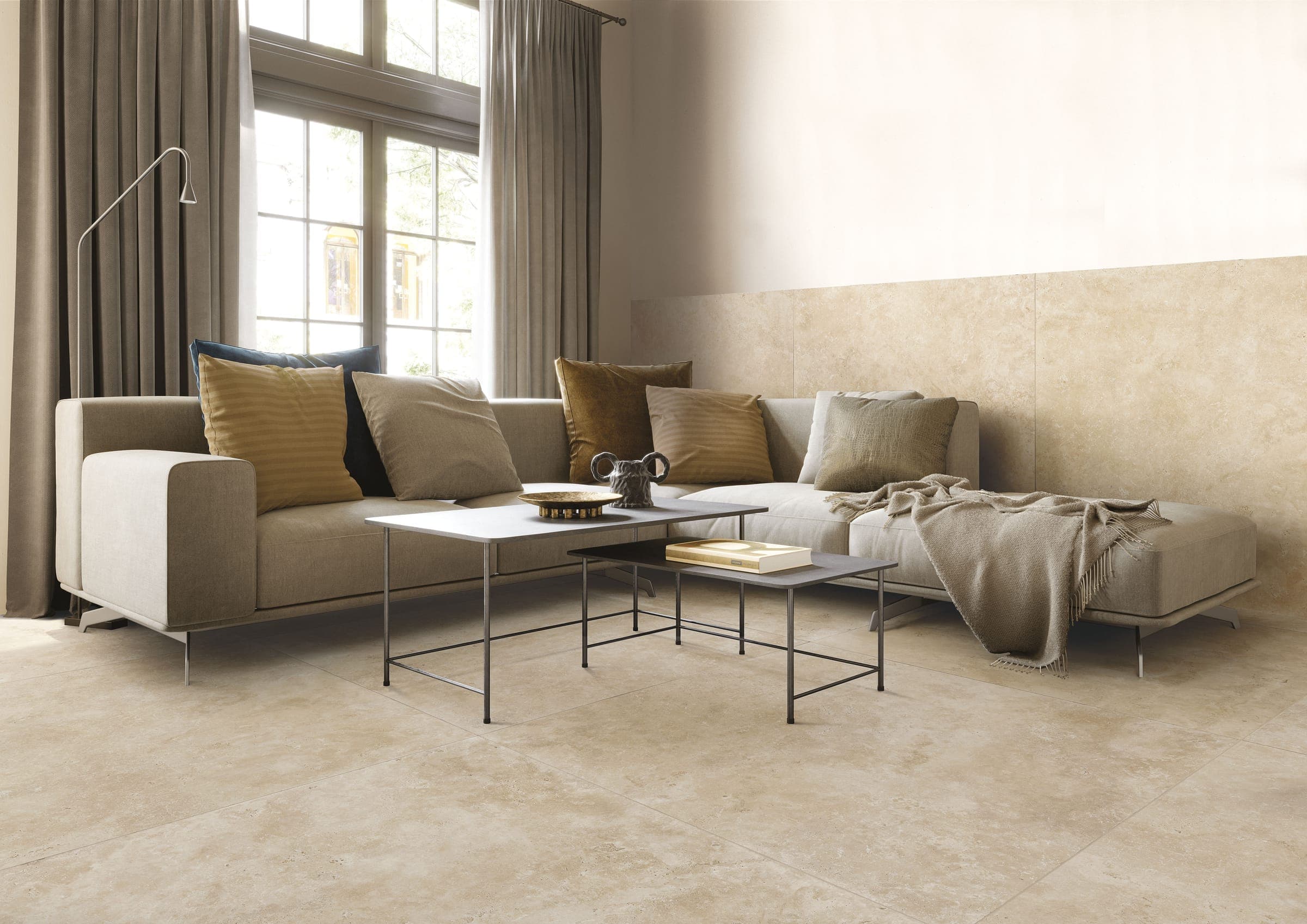Sala con un sofá esquinero gris claro con cojines mostaza, cafés y grises, dos mesas de centro de metal y una manta; el piso y la parte baja del muro están revestidos con piezas cuadradas grandes color beige con aspecto de piedra, junto a un ventanal con cortinas.